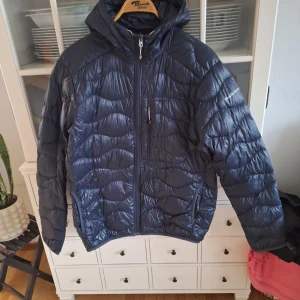 Mörkblå lätt dunjacka från Peak Performance - Säljer en snygg mörkblå lätt dunjacka från Peak Performance. Jackan har en quiltad design med dragkedja framtill och en praktisk huva. Perfekt för kyliga dagar och håller dig varm och bekväm. Passar både till vardags och utomhusaktiviteter.