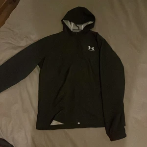 Svart Vår Jacka från Under Armour - Säljer en svart Vår Jacka från Under Armour i storlek M. Den har en luva och är tillverkad i ett syntetmaterial. Perfekt för träning eller vardagsbruk. Den har en lös passform för extra komfort.