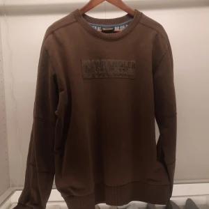 Brun sweatshirt från Wrangler - Säljer en brun sweatshirt från Wrangler med ett tryck på bröstet. Tröjan har långa ärmar och ribbade muddar vid ärmslut och nederkant. Perfekt för en avslappnad stil.