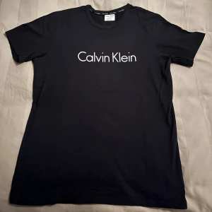 Svart t-shirt från Calvin Klein - Snygg svart t-shirt från Calvin Klein med vit logga på bröstet. Perfekt för en stilren look. Kortärmad och tillverkad i mjukt material för extra komfort.