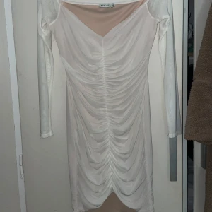 Vit långärmad klänning från NLY One - Snygg vit klänning från NLY One med lång ärm och rynkad design framtill. Klänningen har en v-ringad hals och är tillverkad i ett transparent material som ger en elegant look. Perfekt för en stilren outfit.