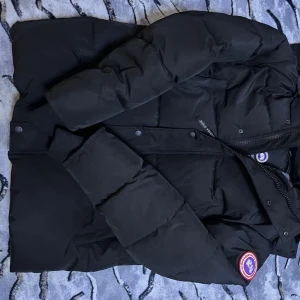 Svart dunjacka från Canada Goose - Säljer en svart dunjacka från Canada Goose med deras ikoniska märke på ärmen. Jackan har långa ärmar och stängs med dragkedja och knappar. Perfekt för kalla dagar.