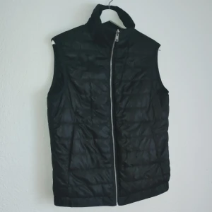 Svart dunväst från J.Lindeberg - Snygg svart dunväst från J.Lindeberg med dragkedja framtill. Perfekt för lager-på-lager under kyliga dagar. Västen har en stilren design och är lätt att matcha med olika outfits.