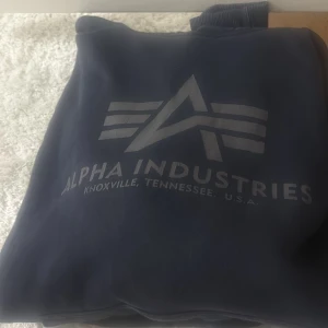 Mörkblå hoodie från Alpha Industries - Säljer en snygg mörkblå hoodie från Alpha Industries med deras ikoniska logga tryckt på framsidan. Hoodien har en klassisk design med dragsko i huvan och en praktisk magficka. Perfekt för en avslappnad stil. 🧢