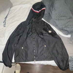 Svart vindjacka med huva - Säljer en snygg svart vindjacka från Moncler med huva och dragkedja. Jackan har en stilren design med röd och vit randdetalj på huvan. Perfekt för blåsiga dagar. Jackan är i storlek 2 den är helt ny aldrig använt 