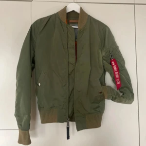 Grön bomberjacka från Alpha Industries - Säljer en grön bomberjacka från Alpha Industries med den klassiska 'Remove Before Flight' taggen. Jackan har en dragkedja framtill och ribbade muddar vid ärmslut och nederkant. Insidan är i en kontrasterande orange färg. Perfekt för en cool och avslappnad stil.