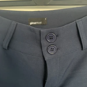 Mörkblå byxor från Gina Tricot - Snygga mörkblå byxor från Gina Tricot med bootcut-stil. Endast använda en gång. Byxorna har mellan hög midja och knappar framtill samt fickor med knappdetalj bak. Perfekta för en stilren look.
