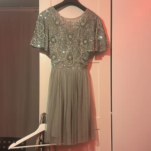 Grön paljett klänning med öppen rygg - Säljer en fantastisk grön klänning med glittriga detaljer och öppen rygg. Klänningen har korta ärmar och en vacker, flödande kjol. Perfekt för en speciell kväll!