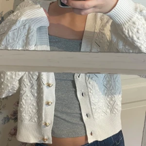 Vit stickad kofta med knappar - Säljer en söt vit stickad kofta med dekorativa knappar. Koftan har ett fint mönster och långa ärmar, perfekt för att hålla sig varm och stilren. Passar bra till både jeans och kjol. Perfekt för höst och vår!