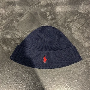 Mörkblå mössa från Polo Ralph Lauren - Snygg stilren mörkblå mössa från Polo Ralph Lauren med röd broderad logga framtill. Skick 9/10 som ny utan prislapp bara och nypris ligger på 799kr! Den är stickad och har en ribbad kant som ger en skön passform. Perfekt för kyliga dagar! 