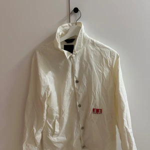Vit jacka från J.Lindeberg - Säljer en stilren vit jacka från J.Lindeberg med knappar framtill och ett tryck på ryggen. Jackan har en hög krage och ett rött JL-märke på bröstet. Perfekt för en cool och avslappnad look.