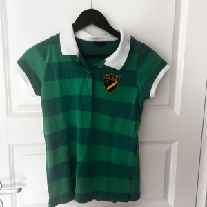 Grön randig pikétröja från GANT - Säljer en snygg grön randig pikétröja från GANT med vit krage och ärmslut. Tröjan har en broderad logga på bröstet och är perfekt för en avslappnad stil. Passar bra till både jeans och shorts. Storlek L men passar mig som s 