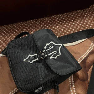 Svart trapstar väska  - Snygg svart axelväska från Trapstar med ett unikt spänne framtill. Väskan har en justerbar axelrem och är perfekt för att bära dina nödvändigheter. Den har en stilren design med ett diskret mönster och märkeslogga.