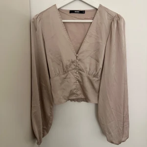  blus  - Snygg beige blus från Bershka med v-ringning och knappar framtill. Blusen har långa ärmar och en smockdetalj i midjan som ger en fin passform. Perfekt för en stilren look.