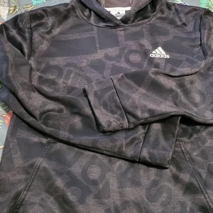 Svart hoodie från Adidas - Säljer en svart hoodie från Adidas med diskret mönster och logga på bröstet. Tröjan har en stor ficka framtill och är tillverkad i mjukt material med fleecefoder. Hund finns hemma.
