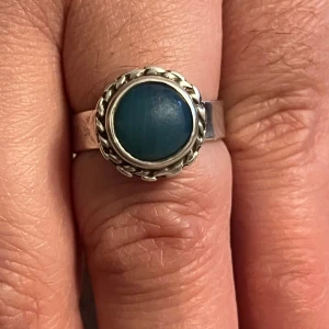 Vintage Silverring 925  med blå sten - Elegant silverring med en rund blå sten i mitten, omgiven av en flätad detalj. Ringen har stämplar på insidan som indikerar äkthet och kvalitet. Perfekt för att ge en stilfull touch till din look.