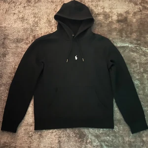 Svart hoodie från Ralph Lauren - Säljer en stilren svart hoodie från Ralph Lauren med det klassiska logotypbroderiet på bröstet. Tröjan har en känguruficka och justerbar huva med snören. Perfekt för en avslappnad look.