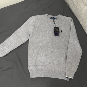 Grå tröja från Polo Ralph Lauren - Snygg grå tröja från Polo Ralph Lauren med klassisk design. Tröjan har långa ärmar och en liten broderad logotyp på bröstet. Perfekt för en stilren look. Helt ny!!!