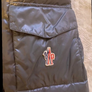 Grå pufferjacka från Moncler grenoble - Säljer en snygg svart pufferjacka från Moncler med broderad logga på ärmen. Jackan har en dragkedja framtill och praktiska fickor. Perfekt för kyliga dagar med sin vadderade design och huva. Skick 7. Storlek 3/M L