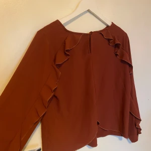 Amber blus med volangdetaljer - Säljer en snygg vinröd/orange/brun blus med volangdetaljer längs ärmarna. Blusen har långa ärmar och en rund halsringning. Perfekt för en stilren look. OBS första bilden är baksidan, det är baksidan som har volanger.