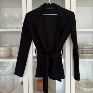 Svart kavaj/blazer med knytband - Bra skick. Svart mjuk blazer. Passar storlek 34 bra. Kommer ej ihåg vart ja. Köpte den, men tror att hemsidan hette något på rebellious.