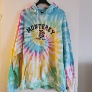Flerfärgad tie-dye hoodie med Monterey-tryck - Säljer en färgglad tie-dye hoodie med texten 'Monterey California' på framsidan. Hoodien har en klassisk passform med långa ärmar och en stor ficka framtill. Perfekt för en avslappnad stil med en touch av färg. Tillverkad i en bomulls- och polyesterblandning. Köpt i Monterey bay, Californien. 