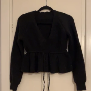 Svart stickad kofta med knytband - Säljer en svart stickad kofta med knytband i midjan och volangdetaljer. Koftan har långa ärmar och en v-ringad design. Perfekt för att hålla sig varm och stilren. Från SHEIN. 