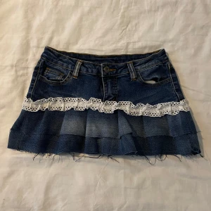 Jeanskjol med spetsdetalj - Snygg jeanskjol med en unik spetsdetalj runt midjan. Kjolen har en kort längd och fransiga kanter för en trendig look. Perfekt för att ge din outfit en cool och avslappnad stil. Midjemått ca 37cm tvärs över.