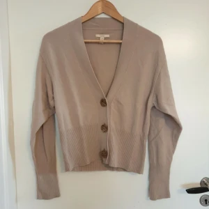 Beige kashmirkofta med knappar - Säljer en elegant beige kashmirkofta med tre stora knappar framtill. Koftan har en djup v-ringning och långa ärmar med ribbade muddar. Perfekt för en stilren och bekväm look.