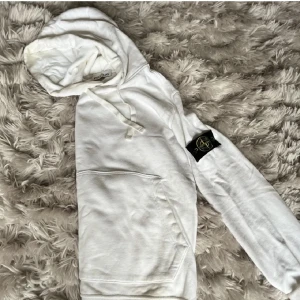 Vit hoodie från Stone Island - Säljer en stilren vit hoodie från Stone Island med klassisk logga på ärmen. Tröjan har en bekväm passform med känguruficka och justerbar huva. Perfekt för en avslappnad look.