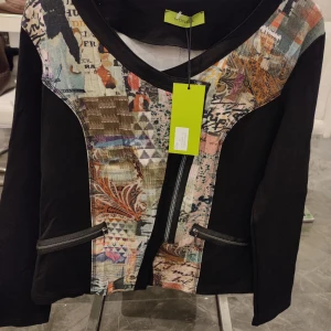 Svart jacka med färgglatt mönster från Mongul - Säljer en unik svart jacka från Mongul med ett färgglatt och konstnärligt mönster framtill och baktill. Jackan har en dragkedja och två fickor med läderdetaljer. Perfekt för dig som vill sticka ut med stil! 🖤