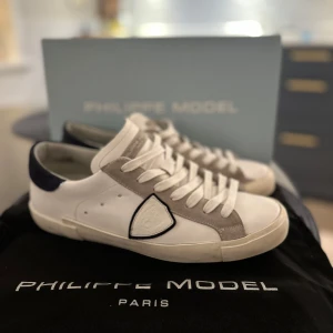 Philippe Model - Säljer ett par snygga vita sneakers från Philippe Model i storlek 41. Skorna har en stilren design med gråa mockadetaljer och mörkblå häl. De är i mycket bra skick. Box och dustbag medföljer. Hör av vid frågor eller funderingar. Kan ta byten. Phillipe model är lite stora i storleken så de passar storlek 42 också. 