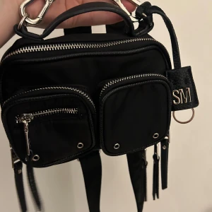 Svart axelväska från Steve Madden - Snygg svart axelväska från Steve Madden med flera praktiska fack och dragkedjor. Väskan har en justerbar axelrem och en liten handtag för enkel bärning. Perfekt för både vardag och fest!