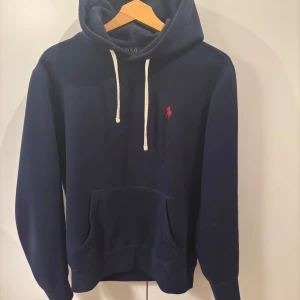 Ralph Lauren Hoodie  - Säljer en snygg mörkblå hoodie från Ralph Lauren med det klassiska röda logotypen på bröstet. Den har en känguruficka och justerbara snören i huvan. Perfekt för en avslappnad stil eller kyliga dagar. Passar både till vardags och chillkvällar. Priset är inte hugget i sten!