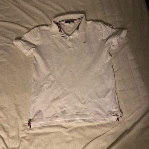 Vit kortärmad pikétröja från Tommy Hilfiger - Säljer en stilren vit pikétröja från Tommy Hilfiger i storlek 164. Tröjan har korta ärmar och klassisk krage med röd och blå randdetalj. Perfekt för både vardag och festliga tillfällen. Skön och bekväm passform!