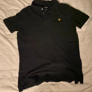 Svart kortärmad piké från Lyle & Scott - Säljer en svart kortärmad pike från Lyle & Scott i bra skick. Den har en klassisk design med en liten gul logga på bröstet och knappar vid kragen. Perfekt för både vardag och festliga tillfällen! Har använt den många gånger och knappat sliten samt så är den extremt skön 