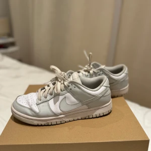 Nike Dunk sneakers i vitt och ljusgrått - Säljer ett par Nike Dunk sneakers i vitt och ljusgrått. De har en klassisk design med snörning och är i bra skick. Perfekta för vardagsbruk eller en avslappnad stil. Storlek är inte specificerad, men de ser ut att passa de flesta.