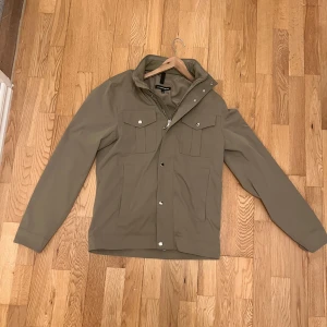 Beige jacka med dragkedja - Säljer en snygg beige jacka i mycket bra skick. Den har en dragkedja och knappar framtill samt två bröstfickor med lock. Perfekt för höst och vår, med en stilren och enkel design. Jackan är långärmad och passar bra till både vardag och lite finare tillfällen.