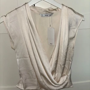 Beige omlottopp från Mango  - Snygg omlottopp i satin från Mango. Toppen har en draperad design framtill och är ärmlös, i en krämvit/vit färg. Materialet är mjukt och bekvämt. Går att använda både till vardags och om man ska klä upp sig🩷