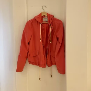 Korallfärgad jacka från ONLY - Säljer en snygg korallfärgad jacka från ONLY i storlek S. Jackan har en huva med dragsko och praktiska fickor framtill. Perfekt för vår och sommar med sin lätta design. Den har långa ärmar och stängs med dragkedja och knappar. Passar både till vardags och lite finare tillfällen.