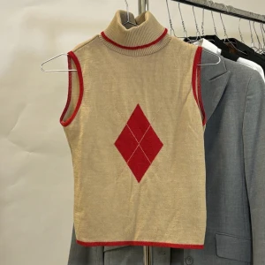 Beige ärmlös topp med röd diamant - Snygg beige ärmlös topp med en röd diamantformad detalj på framsidan. Den har en hög krage med röda kanter som matchar de röda detaljerna vid ärmhålen och nederkanten. Perfekt för en stilren och retro look!