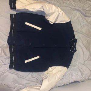 Blå och vit bomberjacka - Säljer en snygg blå och vit bomberjacka med långa ärmar. Jackan har knappar framtill och vita detaljer på fickorna. Perfekt för höst och vår! 🧥