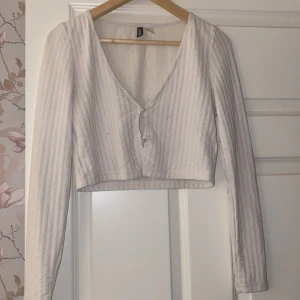 Vit ribbad croppad topp från H&M Divided - Säljer en superfin vit ribbad topp från H&M Divided. Den är croppad med en söt knytning framtill och har långa ärmar. Perfekt för både vardag och fest! Toppen är i bra skick med några små tecken på användning.