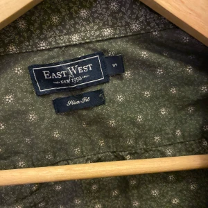 Grön skjorta från East West - Säljer en snygg grön skjorta från East West i storlek S. Skjortan har ett diskret blommigt mönster och är i slim fit. Perfekt för både vardag och festliga tillfällen. Den är i mycket bra skick och redo för en ny ägare!