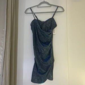 Glittrig blå klänning - Snygg glittrig blå klänning med smala axelband och draperad design. Perfekt för festliga tillfällen! Den är kort och har en figurnära passform som framhäver formerna. Materialet är glittrigt och ger en lyxig känsla.