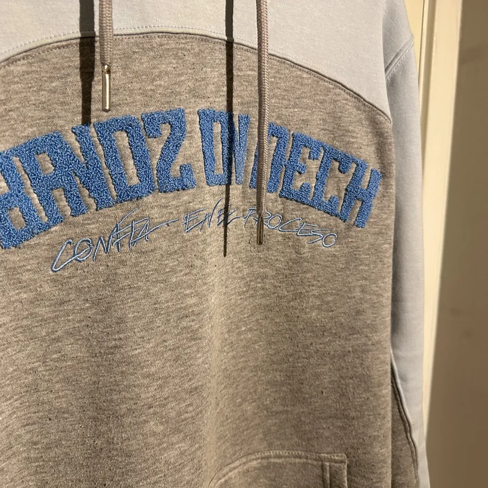 Säljer en snygg grå och vit hoodie med blå text på bröstet. Den har en stor ficka fram och en bekväm huva. Perfekt för en avslappnad stil och passar bra till jeans eller joggers. Skön att ha på sig under kyliga dagar.. Tröjor & Koftor.