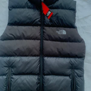 Svart dunväst från The North Face - Säljer en snygg och varm svart dunväst från The North Face. Perfekt för kyliga dagar med sin quiltade design och dragkedja framtill. Västen har en normal passform och är i nytt skick. Idealisk för höst och vinter! Västen är splitter ny med lapp och kvitto.Köpt på Londons Flyhplats för ett superpris därav varför jag säljer