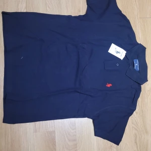 Svart pikétröja från Polo Sport - Säljer en klassisk svart pikétröja från Polo Sport i 100% bomull. Den har en broderad röd logga på bröstet och knappar vid kragen. Perfekt för en stilren och avslappnad look!