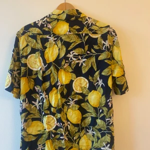 Mönstrad kortärmad skjorta med citroner - Säljer en supercool kortärmad skjorta med ett färgglatt citronmönster. Perfekt för sommaren! Skjortan har en mörkblå bas med stora gula citroner och gröna blad. Den är i bra skick och har knappar framtill. Perfekt för en somrig fest eller bara för att liva upp vardagen! 🍋