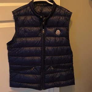 Blå dunväst från Moncler - Säljer en snygg blå dunväst från Moncler i rätt bra skick. Det finns ett litet hål på baksidan som knappt syns. Västen har en dragkedja framtill och två praktiska fickor med dragkedja. Passar både till vardags och lite finare tillfällen. Kan gå ner i pris vid snabb affär! Hör av er vid fler frågor och bilder!